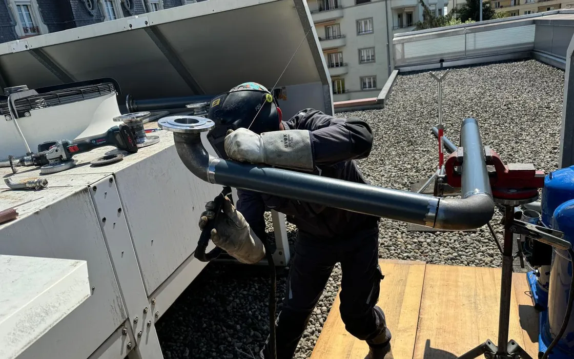 Un soudeur en masque raccorde une conduite bridée à une pompe à chaleur air-air installée sur un toit plat, devant des façades d’immeubles lausannois.
